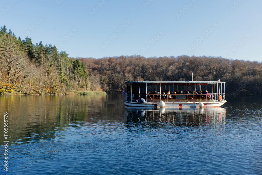 Fototapeta premium Plitvice Lake, surrounded by vibrant autumn forests, offers a serene boat ride with tranquil reflections on its clear waters.