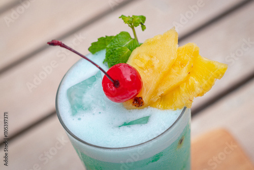 Close up view, Blue hawaii in glass garnished with slice pineapples and cherry and mint leaves on wooden planks in front of a pool photo