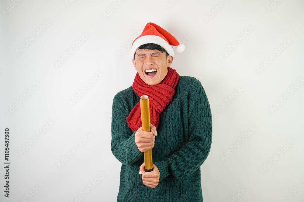 Young asian man wearing santa clause hat, red scarf and green long sleeved sweater with smiling face expression is holding confetti cannon, isolated over white background. Young asian man wearing santa clause hat, red scarf and green long sleeved sweater with smiling face expression is holding confetti cannon, isolated over white background.