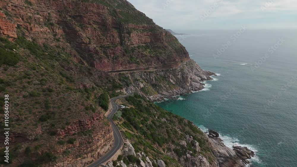 Beautiful cinematic drone shot of a coastal road winding around a mountain covered in flora with a sheer drop to the rocky coastline below Beautiful cinematic drone shot of a coastal road winding around a mountain covered in flora with a sheer drop to the rocky coastline below