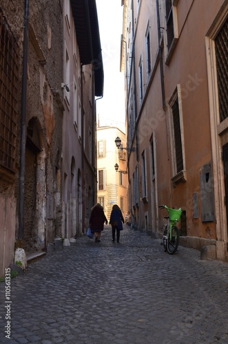 Two people walk along a narrow street between buildings.