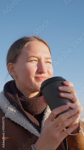 Young woman in warm clothing enjoys drinking takeaway coffee outdoors on sunny autumn day. Bright sunlight enhances her smile as she relaxes in casual urban setting. vertical video