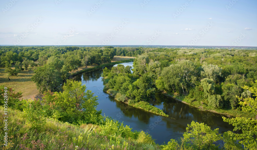Aerial view of a calm river winding through dense green forests and open fields on a clear summer day with blue skies. Aerial view of a calm river winding through dense green forests and open fields on a clear summer day with blue skies.