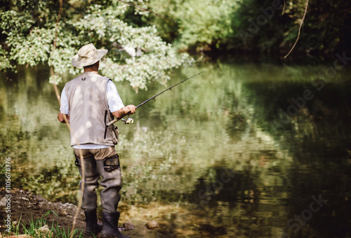 A fisherman standing by a calm river, wearing a fishing vest and hat, holding a fishing rod surrounded by lush green nature. photo