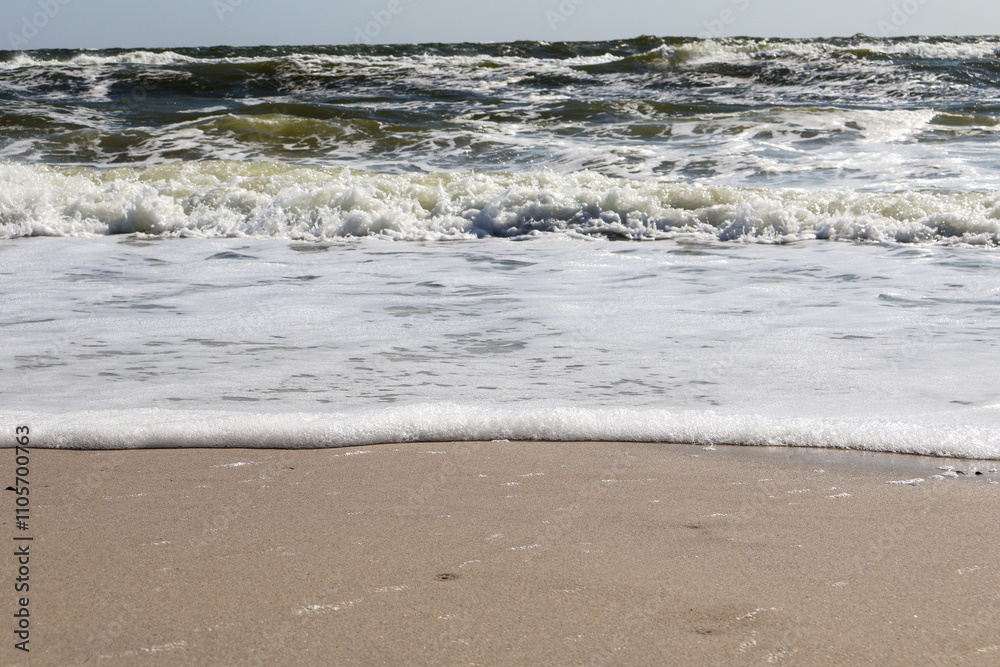 Close-up view of ocean waves creating a foamy edge as they gently reach the sandy shore under sunlight. Close-up view of ocean waves creating a foamy edge as they gently reach the sandy shore under sunlight.