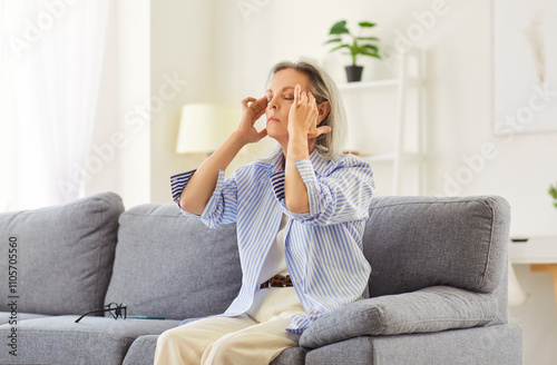 Sick mature woman sitting on sofa, suffering from headache or hypertension, rubbing her temples. Tired pensioner with high blood pressure feeling exhausted and unwell. Cardiovascular diseases concept.