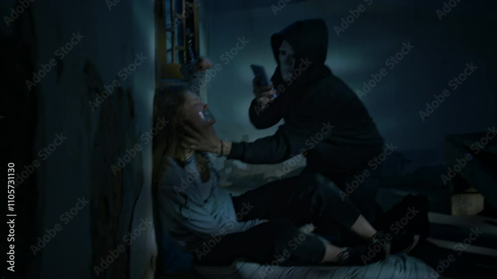 Full side handheld shot of masked perverse criminal taking pictures on smartphone of young poor and desperate female hostage with duct tape on mouth, being handcuffed to window grate in basement Full side handheld shot of masked perverse criminal taking pictures on smartphone of young poor and desperate female hostage with duct tape on mouth, being handcuffed to window grate in basement