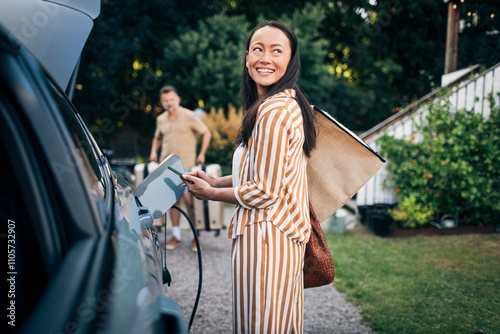 Smiling woman carrying bags while standing near car charging in back yard