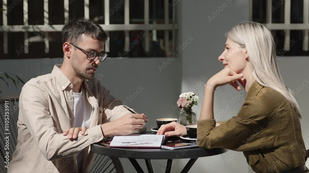 A man and a woman are sitting outside at a cafe, working. On the table there is coffee and a notebook with notes. The business partners are meeting about a project. A man and a woman are sitting outside at a cafe, working. On the table there is coffee and a notebook with notes. The business partners are meeting about a project.