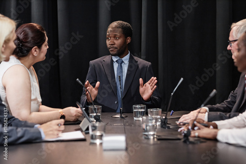 Medium shot of confident African American male presidential candidate speaking with political party committee developing campaign strategy while sitting at table against black curtains, copy space photo