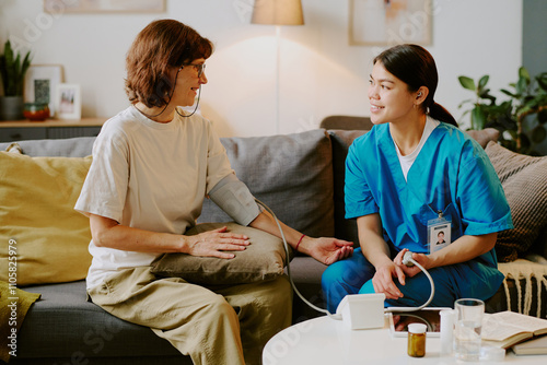 Elderly person having health checkup from smiling nurse in living room, nurse wearing scrubs measuring blood pressure while engaging in conversation