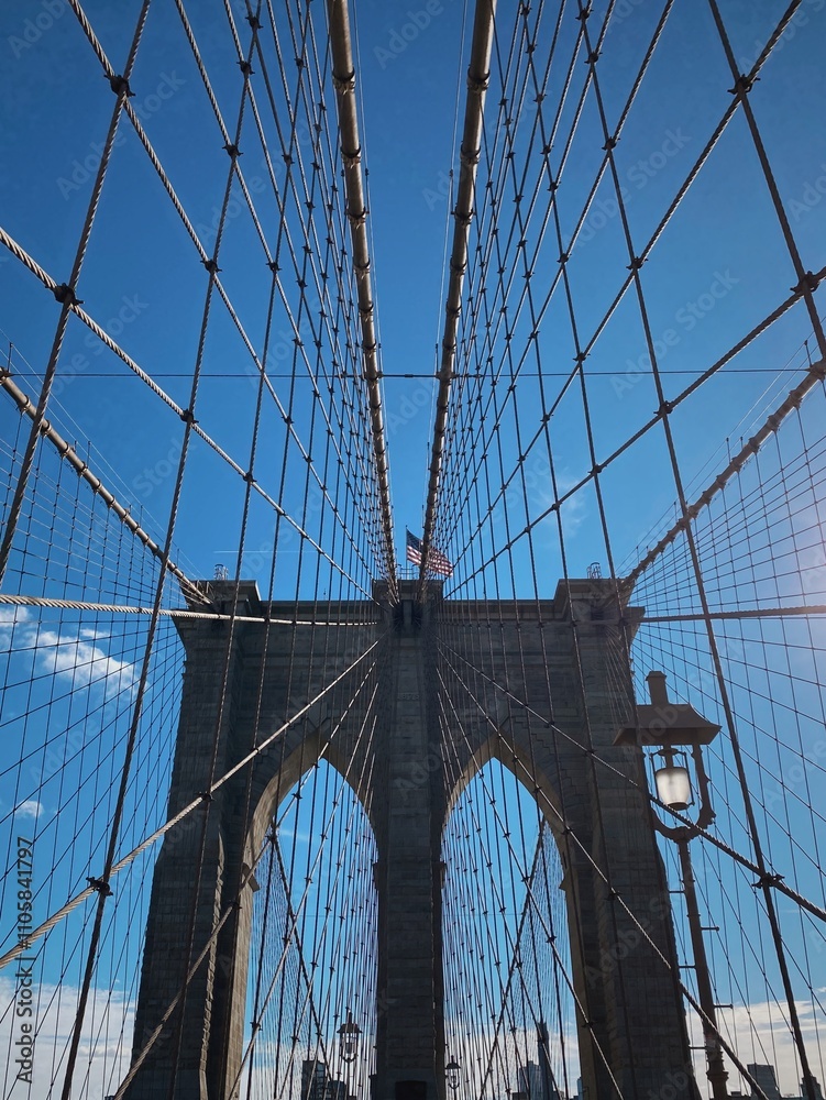Obraz premium A sunny day walk across the iconic Brooklyn Bridge in New York City, showcasing its intricate suspension cables and stunning architectural details against a clear blue sky