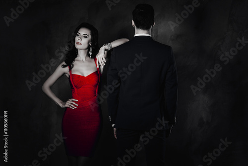 Portrait of brunette lady with curly hair in violet dress with bijoutery looking to the side holding hand on waist lean on shoulder of latin gigolo man with rear back view isolated on grey background photo