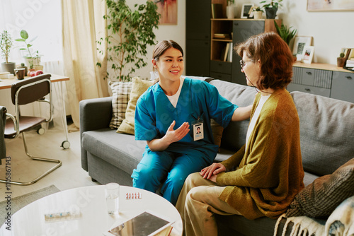 Healthcare worker sitting beside elderly lady in cozy living room providing comfort and care, indoor setting with natural light filtering through windows