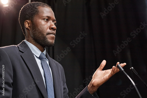 Portrait shot of adult African American male politician in blue suit gesticulating with hands addressing audience while giving public speech on stage in spotlight against black curtains, copy space photo