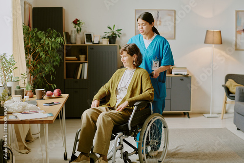 Nurse providing support for senior woman in wheelchair in cozy living room with plants and furniture in background photo