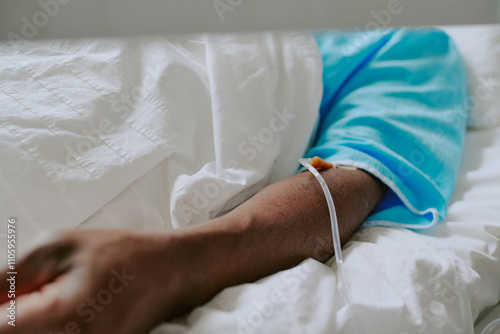 Patient lying in hospital bed with IV drip connected into arm, foreground showing close-up of patients arm and medical equipment used for treatment