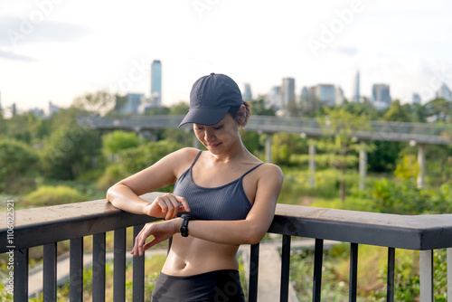 Asian woman lean wall railing on walkway bridge and look at her watch to relax after exercise in public garden with warm light in evening.