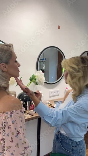 Professional make-up artist finishes doing make-up for a beautiful blonde girl. Make-up with red lipstick. Beautiful studio, lots of cosmetics, reflection in the mirror.