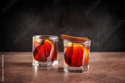 Two glasses of homemade traditional winter red mulled wine drink or grog cocktail made with slices of fresh orange citrus fruit decorated with cinnamon stick served on brown stony table on black wall