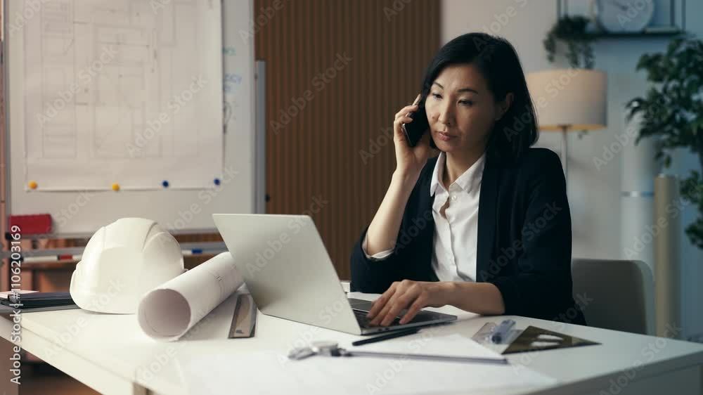 Middle-aged Asian female architect talks on phone with a contractor in office Middle-aged Asian female architect talks on phone with a contractor in office