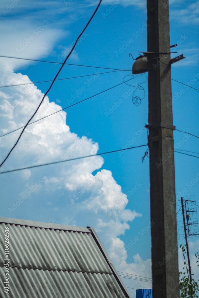 a bright blue sky filled with large, fluffy white clouds. In the foreground, there is a utility pole with several wires extending from it a bright blue sky filled with large, fluffy white clouds. In the foreground, there is a utility pole with several wires extending from it