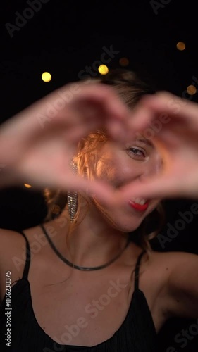 Winter holidays mood. Blonde woman smiling showing heart symbol with hearts. Elegant evening style. Millennial Hand Heart. Dark black background Slow motion vertical video footage