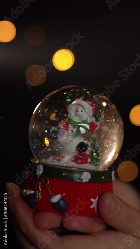 Hands female holding crystal snow ball with funny snowman inside. Dark black background with Christmas lights. Slow motion vertical video footage. 