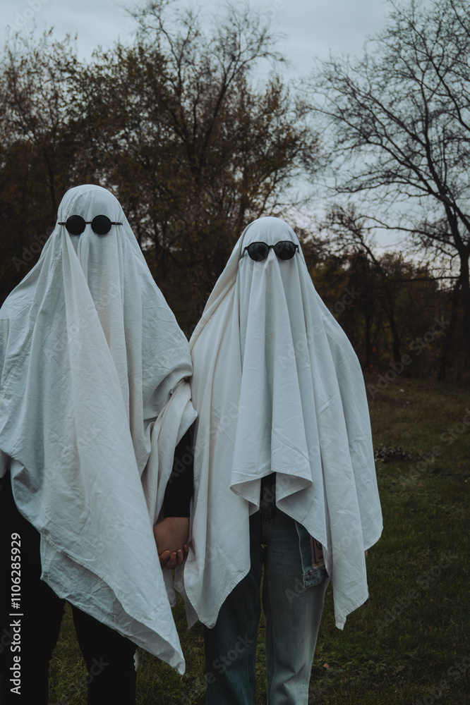 In a moody outdoor setting, two figures draped in white sheets resemble playful ghosts, each wearing dark sunglasses, holding hands against a backdrop of trees. In a moody outdoor setting, two figures draped in white sheets resemble playful ghosts, each wearing dark sunglasses, holding hands against a backdrop of trees.