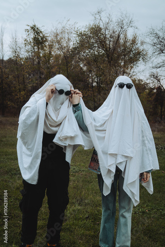 A whimsical scene shows two individuals dressed as ghosts in white sheets, holding hands in a grassy area surrounded by trees, with one wearing dark sunglasses, creating a playful vibe. photo