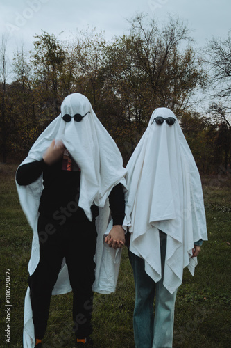 A whimsical scene shows two individuals dressed as ghosts in white sheets, holding hands in a grassy area surrounded by trees, with one wearing dark sunglasses, creating a playful vibe. photo