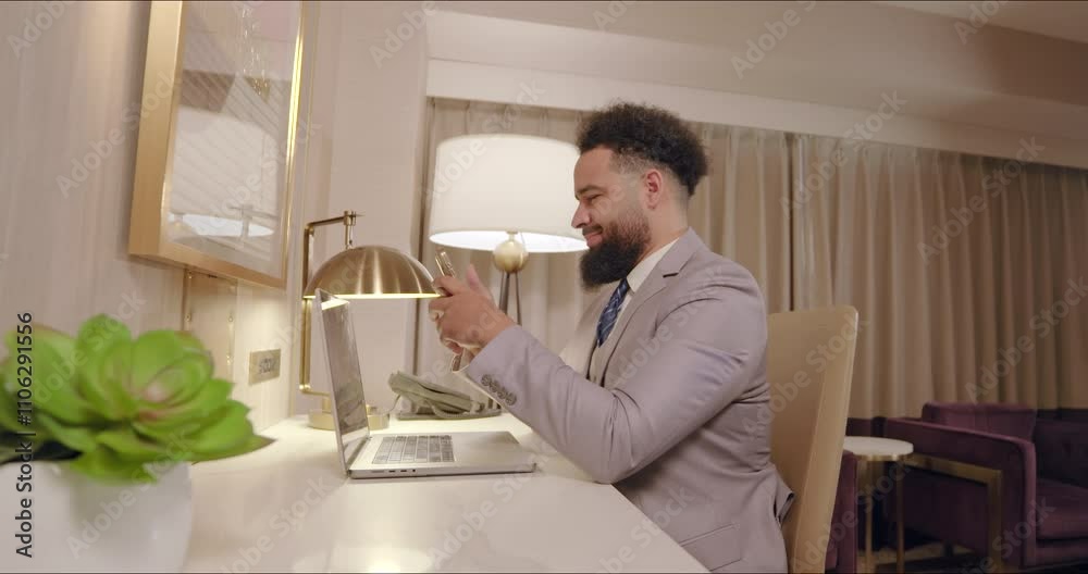Cheerful african american businessman dressed in a sharp suit is fully focused on his phone while simultaneously working on a laptop in a sleek and stylish hotel space, professionalism and modernity Cheerful african american businessman dressed in a sharp suit is fully focused on his phone while simultaneously working on a laptop in a sleek and stylish hotel space, professionalism and modernity