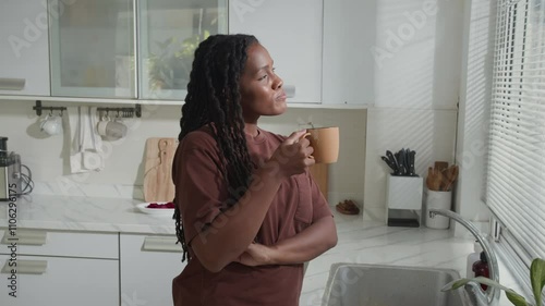 Black young woman drinking coffee and daydreaming while standing near window in kitchen having leisurely morning