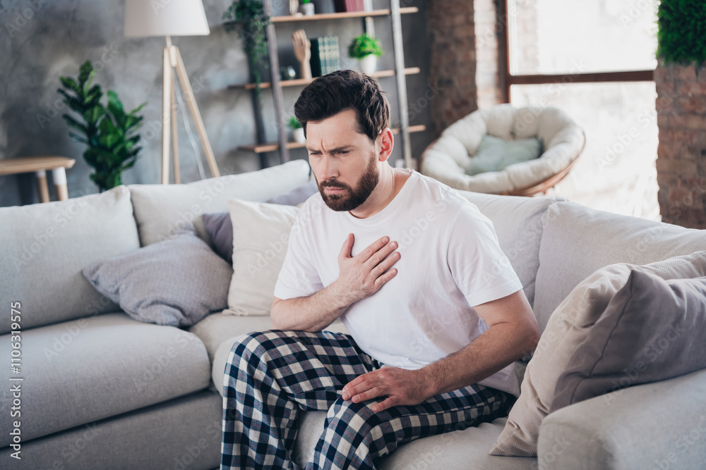 Photo of young man wear pajama sit on sofa arm touch chest heart attack pain hurt living room indoors Photo of young man wear pajama sit on sofa arm touch chest heart attack pain hurt living room indoors