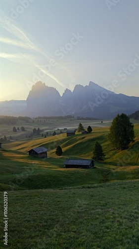 Vertical shot of the Alpe di Siusi at sunset, capturing the Dolomites’ towering peaks and scenic meadow landscape dotted with rustic mountain huts in South Tyrol, Italy.
