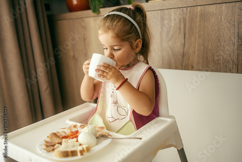 Portrait of a hungry little girl sitting in her chair at home and drinking form mug during breakfast. photo