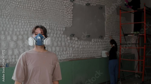 Young decorator, satisfied with completion of her work, removes respirator from her face, which protects her from harmful elements contained in paint. Portrait.
