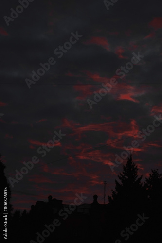 a dramatic sunset with vibrant red and orange hues streaking through a textured sky, silhouetting an old building and tall trees beneath a moody atmosphere