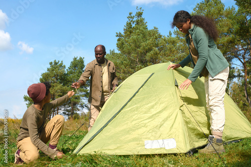 Low angle shot of African American boy sitting on grass and helping his parent and sister with pitching tent in forest while they camping together photo