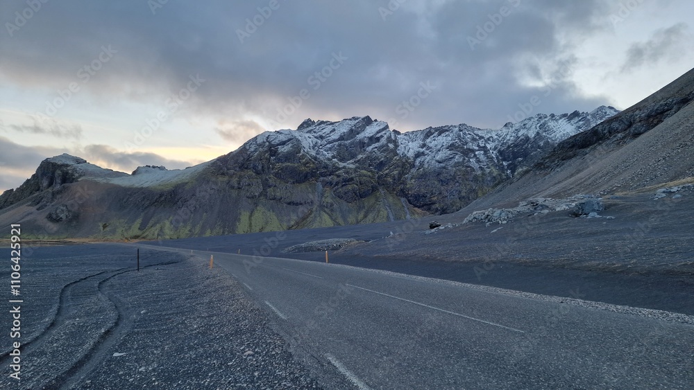 Naklejka premium Snowy mountain cliffs across long road in icelandic nature landscape, highway in wintry weather. Peaks covered in snow alongside lonely road within beautiful nordic valley.