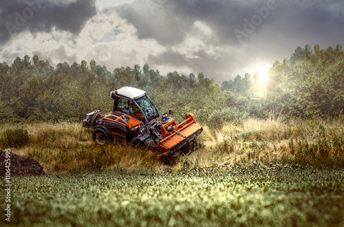 traktor mit rasenmäher schneidet hohes gras an einem hügel mit grünen gras im vordergrund und strahlendem sonnenschein photo