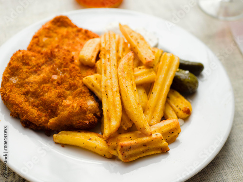 Breaded chicken fillet meal and fried potatoes chips with ridges on a white plate and light color table cloth. Simple food made of high quality premium products. Cooked poultry meat.