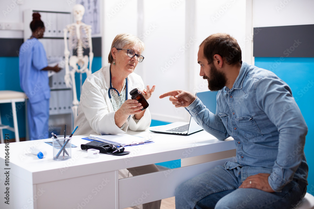Fototapeta premium Friendly older doctor holds a pill bottle while discussing healthcare plan with young white man, guiding him on prescribed medication. Male patient consults with senior physician about his medicine.