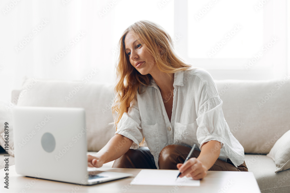 Woman working from home on a cozy couch with laptop and pen, focused on her tasks and taking notes