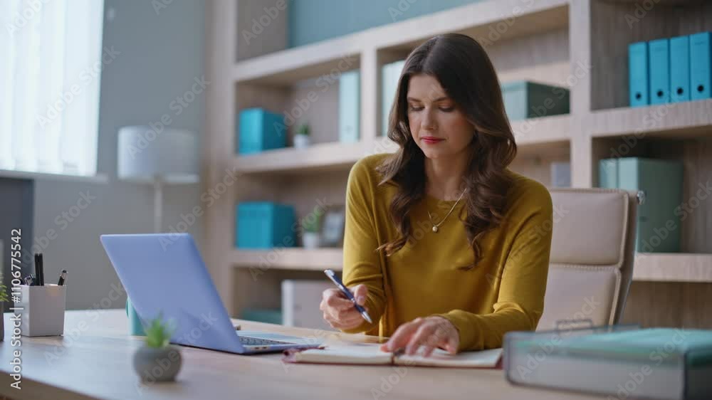 Online student looking laptop noting data at notebook at desk. Successful woman