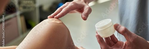 Фотография Hand holding a jar of cream while applying moisturizer on a kneecap in a home environment