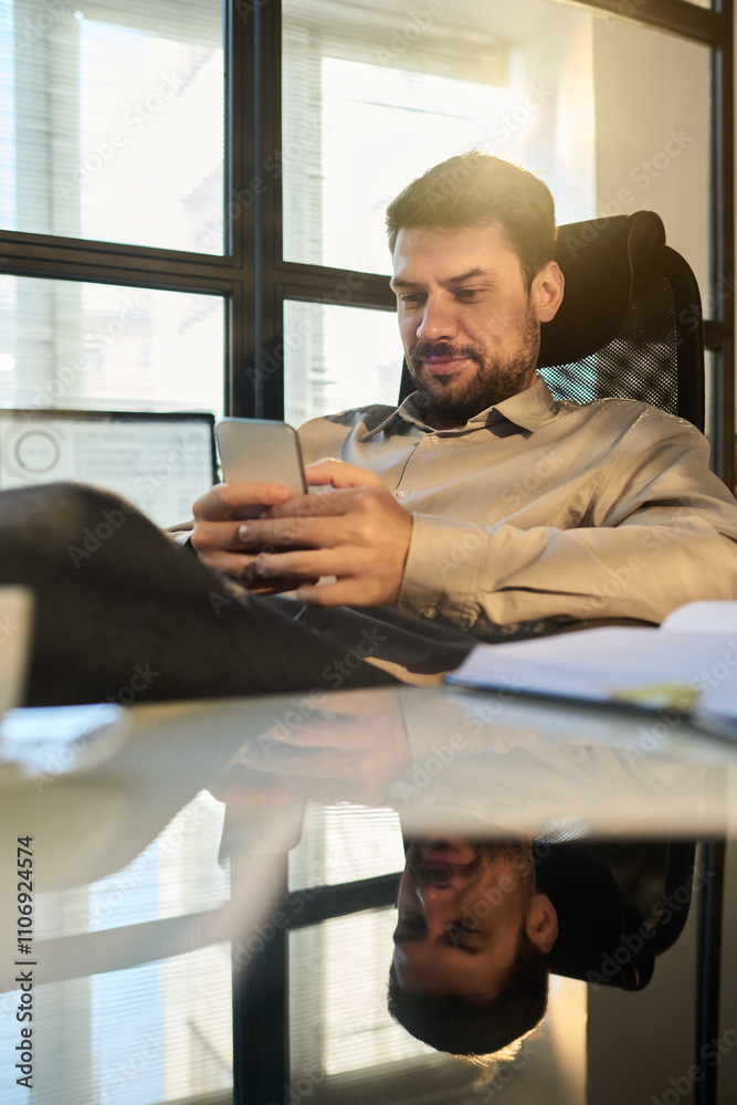 Young restful office manager in smart casualwear texting in smartphone while enjoying break by workplace with notepad Young restful office manager in smart casualwear texting in smartphone while enjoying break by workplace with notepad