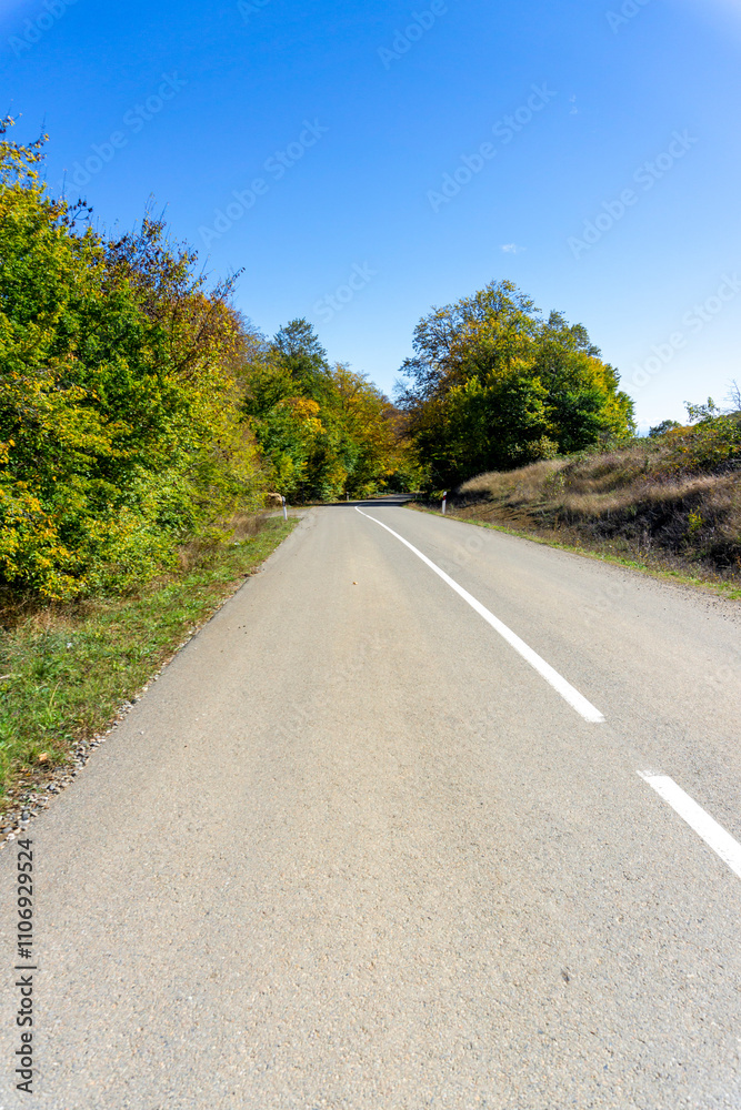 An asphalt road with a white dividing line passes through trees and bushes with autumn leaves. Hills and clouds are in the background An asphalt road with a white dividing line passes through trees and bushes with autumn leaves. Hills and clouds are in the background