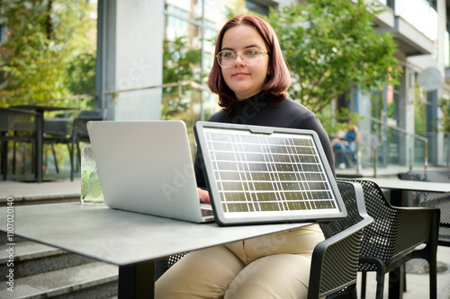 Female using portable solar panel to charge laptop and smartphone. Portrait of a young student girl working remotely from laptop while sitting at a table in a cafe. Bright sunny day. Renewable energy.
