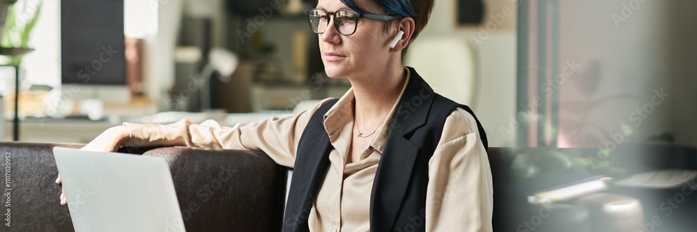 Fototapeta premium Header of young serious female analyst with laptop on her knees sitting on leather couch in office and working with online data